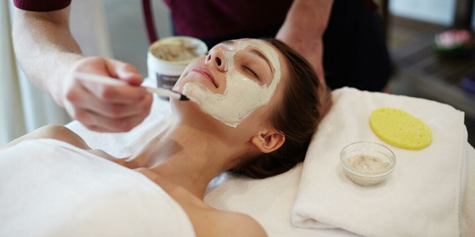 Closeup of unrecognizable male cosmetologist applying face mask with brush to face of beautiful woman lying on massage table in SPA center